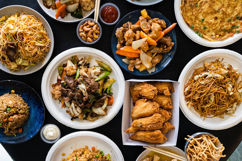 An overhead view of a variety of Chinese takeout dishes on a black table, including fried rice, noodles, orange chicken, beef stir-fry, and fried wings.
