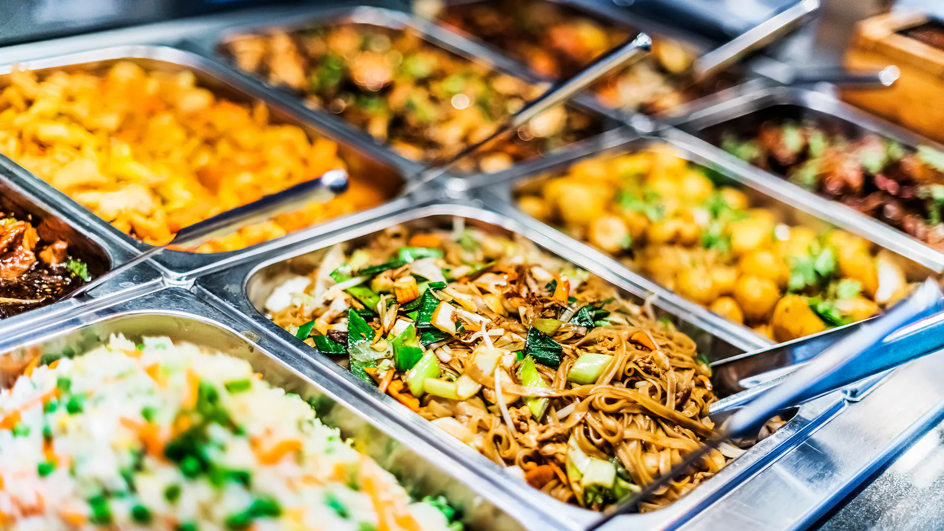 A close-up of a buffet line with stainless steel trays filled with various Asian dishes including fried rice, noodles, and stir-fries.