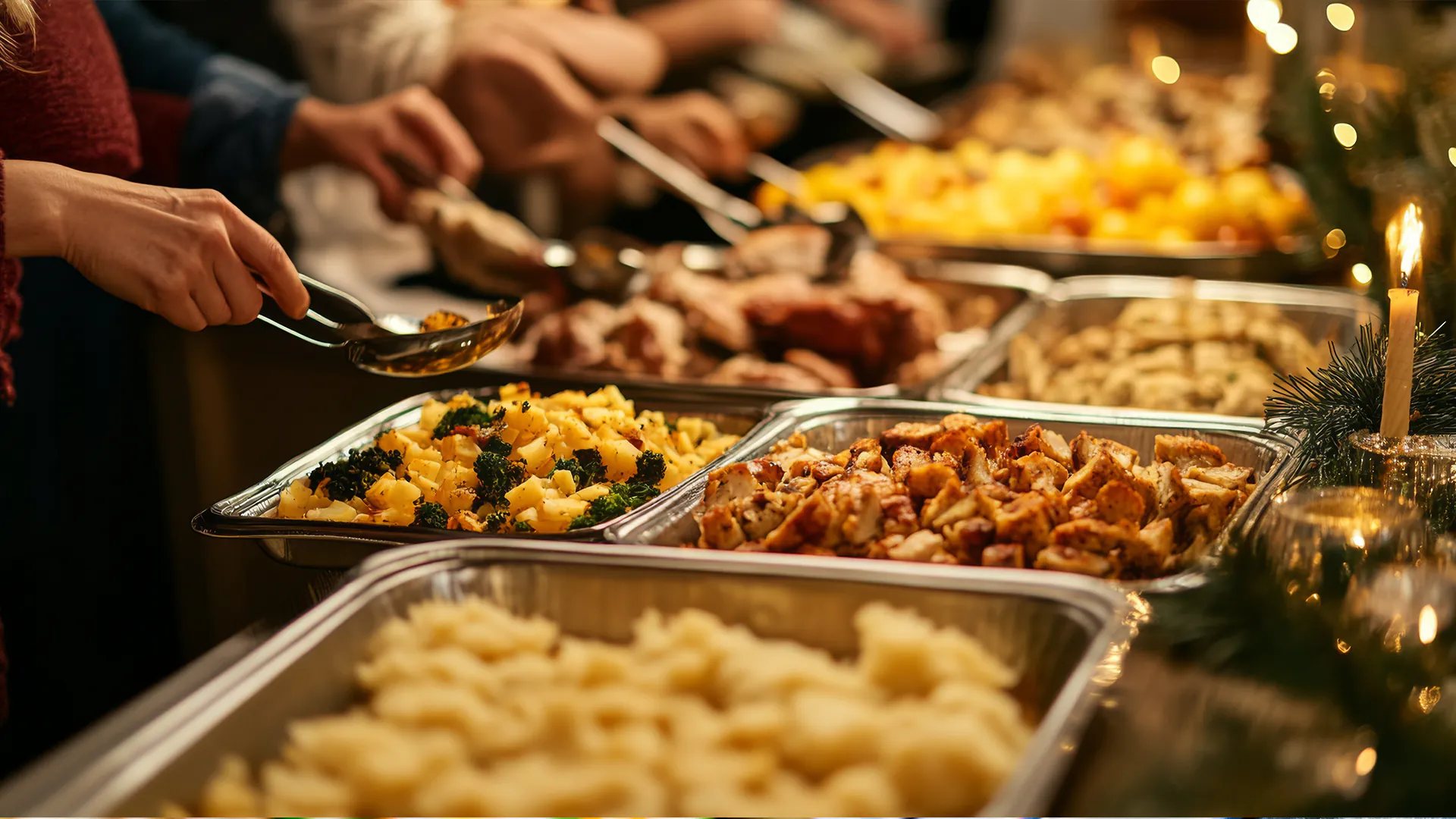 People serving themselves from a festive holiday buffet with warm lighting, candles, and trays of roasted chicken, potatoes, and broccoli.