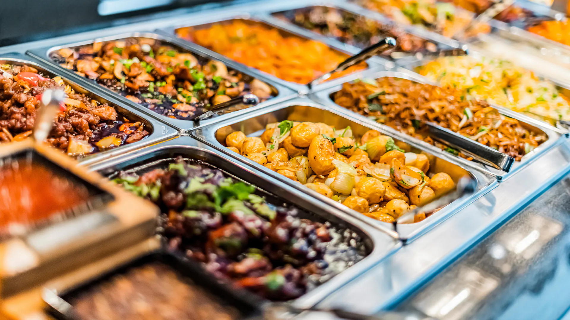 Another angle of the same buffet line, showing more trays of cooked meats, vegetables, and dumplings with serving tongs.